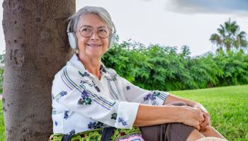 Portrait of relaxed attractive senior woman with headphones sitting in the meadow of public park looking away - caucasian lady enjoying music and free time