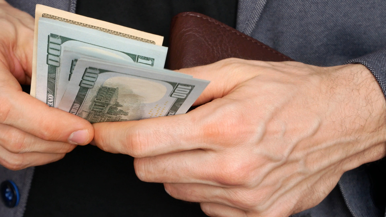 Close-up of the hands of a man in a gray blazer holding a leather wallet and counting out the money