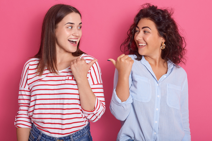 Indoor shot of two young beautiful girls pointing towards each other, brunette women dress shirts, standing against pink studio background, expresses happynes and astonish. Same sex love concept.