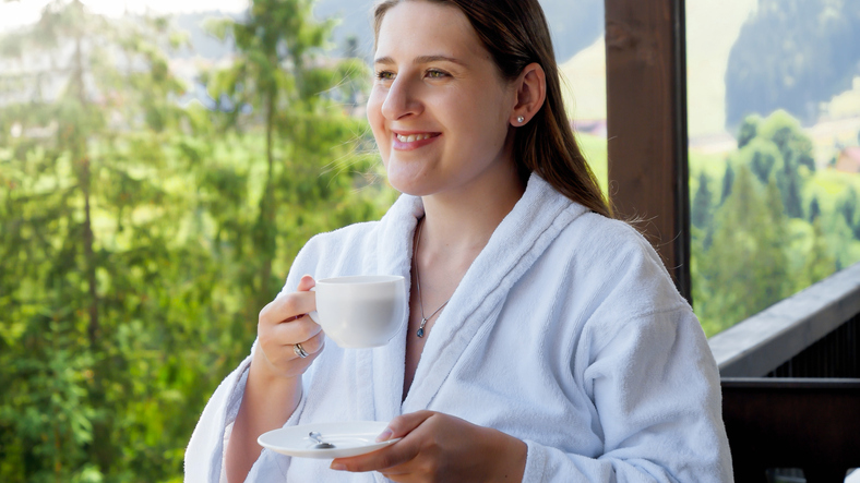 Happy smiling woman in bathrobe drinking coffee and enjoying view on mountains at hotel. Concept of people traveling, tourism and vacation in mountains at summer
